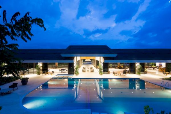 A modern, well-lit building with a large swimming pool in front, surrounded by lounge areas under a twilight sky.