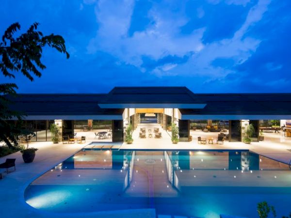 A modern, well-lit building with a large swimming pool in front, surrounded by lounge areas under a twilight sky.