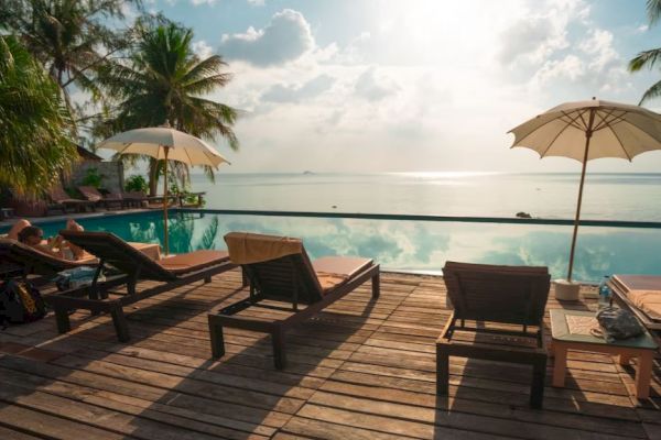 A serene poolside setting with lounge chairs, umbrellas, and palm trees overlooking a calm ocean under a partly cloudy sky.