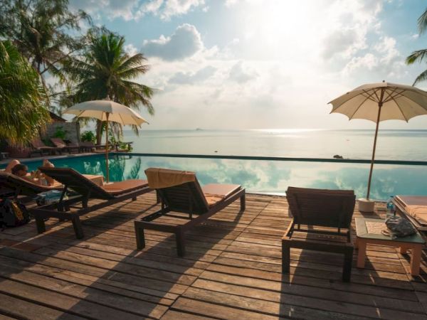 A serene poolside setting with lounge chairs, umbrellas, and palm trees overlooking a calm ocean under a partly cloudy sky.