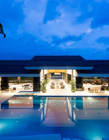 A luxurious pool area with lounge chairs and modern architecture, illuminated in the evening under a blue sky with scattered clouds.