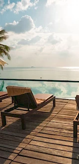 A serene poolside scene with several lounge chairs and umbrellas placed on a wooden deck overlooking a calm water body during the day.