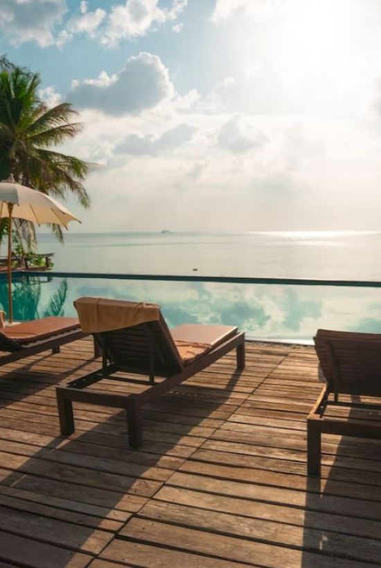 Beachside pool area with lounge chairs and umbrellas, overlooking a calm sea and a clear sky with palm trees around, capturing a serene atmosphere.