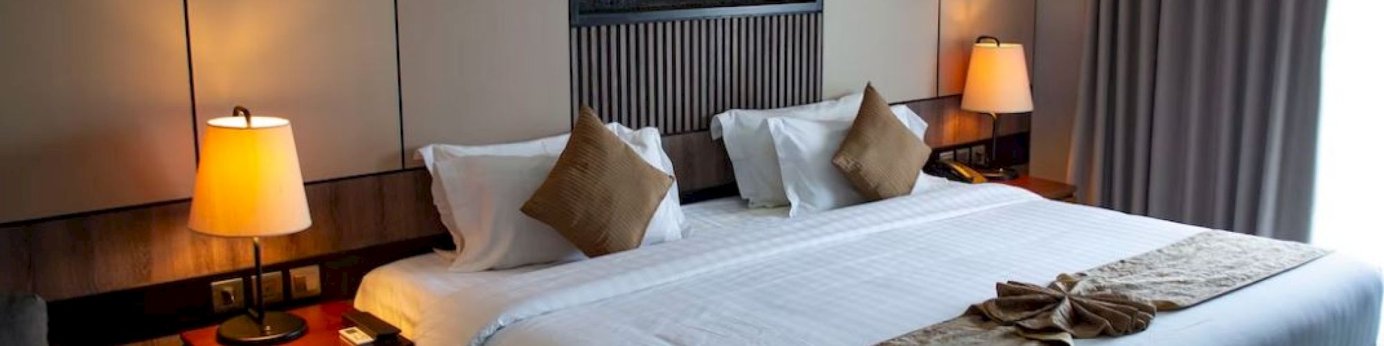 The image shows a neatly made bed with a white duvet, brown accents, and pillows, flanked by two bedside tables with lamps in a cozy, modern room.