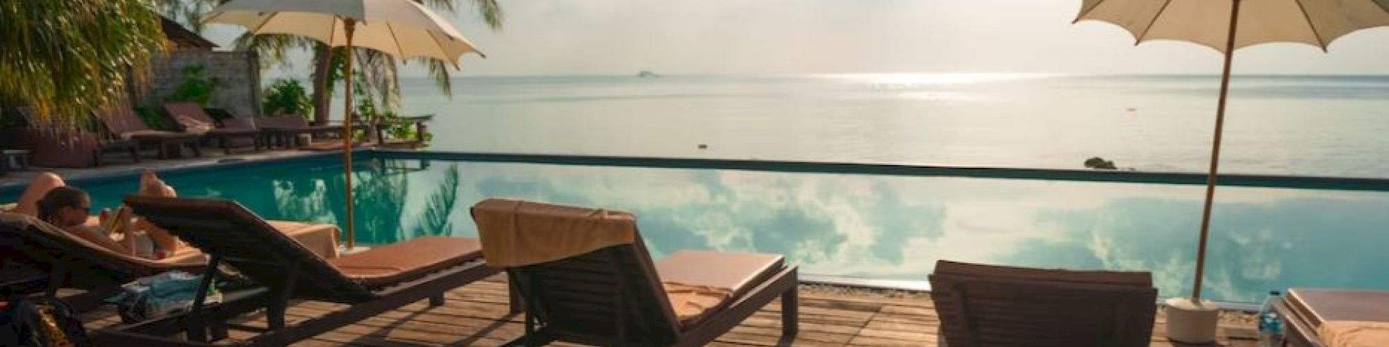 A serene poolside setting with lounge chairs, sun umbrellas, and tropical palm trees, overlooking a tranquil sea under a bright sky.