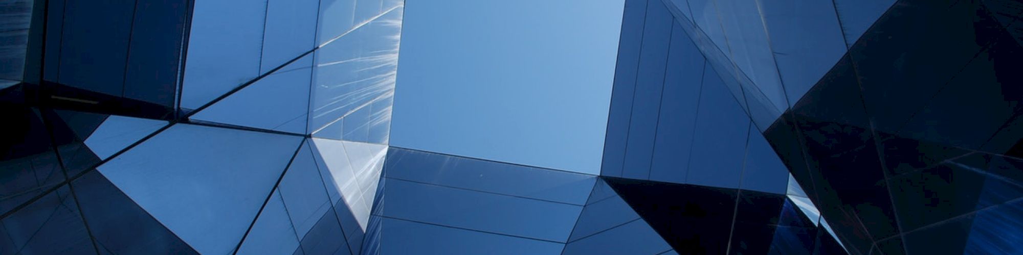 The image shows a geometric perspective looking up through a complex arrangement of reflective glass surfaces, framing the clear blue sky above.