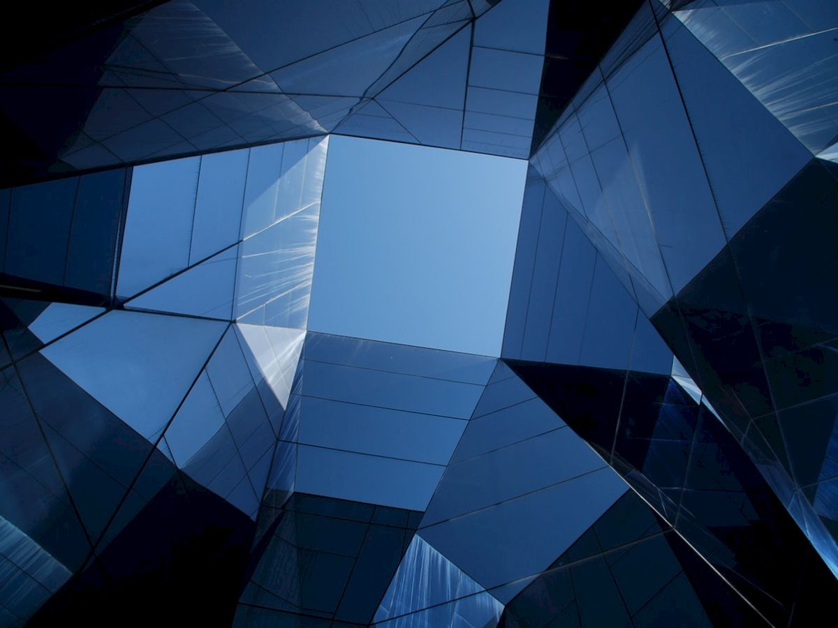 The image features modern, angular glass architecture with reflective surfaces, forming a geometric pattern against a clear blue sky.