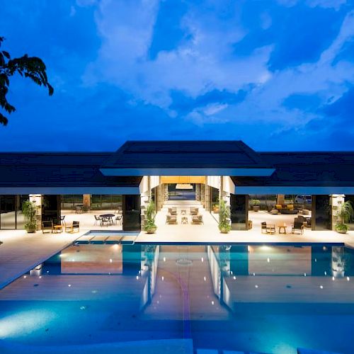 An illuminated pool area at night with a spacious, modern building in the background, featuring deck chairs and outdoor seating.