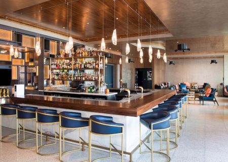 The image shows a modern bar with a wooden countertop, stylish blue chairs, hanging lights, and a well-stocked display of beverages.