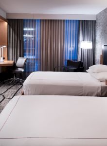 A modern hotel room with two neatly made beds, a TV, a desk and chair, contemporary lighting, and large windows with curtains.