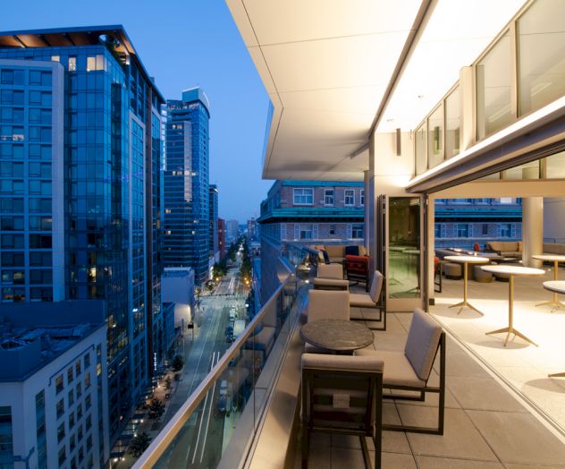 A modern balcony overlooking a city street with skyscrapers. The balcony has seating and tables, and the interior is brightly lit.