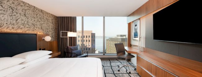 A modern hotel room with a bed, desk, chair, large TV, and a view of buildings and water through large windows, featuring contemporary decor.