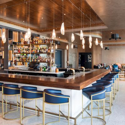 This image shows a modern bar with a polished wooden counter, blue chairs, shelves stocked with bottles, and ambient lighting in a contemporary setting.