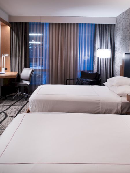 The image shows a modern hotel room with two neatly made beds, a desk, chair, large TV, stylish decor, and ambient lighting.