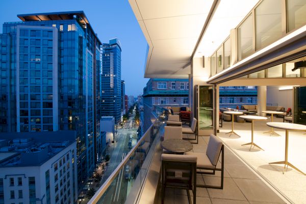 A modern cityscape with tall buildings and a stylish outdoor balcony area with seating and tables, overlooking a well-lit street at dusk.