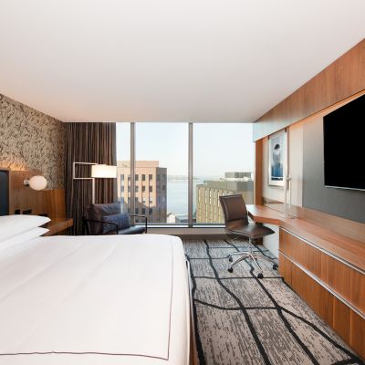A modern hotel room with a large bed, desk, chair, flat-screen TV, abstract-patterned carpet, and a view of the city and water through large windows.
