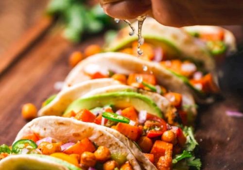 The image shows a hand squeezing lime juice over a row of colorful tacos filled with chickpeas, vegetables, and garnished with herbs.