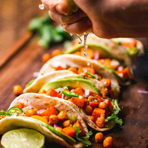 The image shows a hand squeezing lime juice over a row of colorful tacos filled with chickpeas, vegetables, and garnished with herbs.