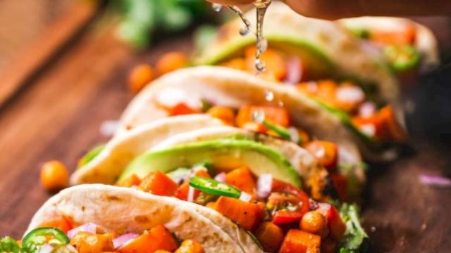 The image shows a hand squeezing lime juice over a row of colorful tacos filled with chickpeas, vegetables, and garnished with herbs.