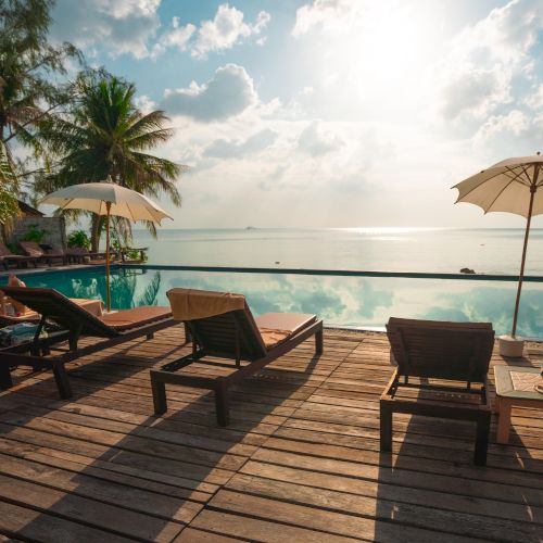 A serene poolside scene with lounge chairs, umbrellas, and palm trees overlooking a tranquil ocean at sunrise or sunset.
