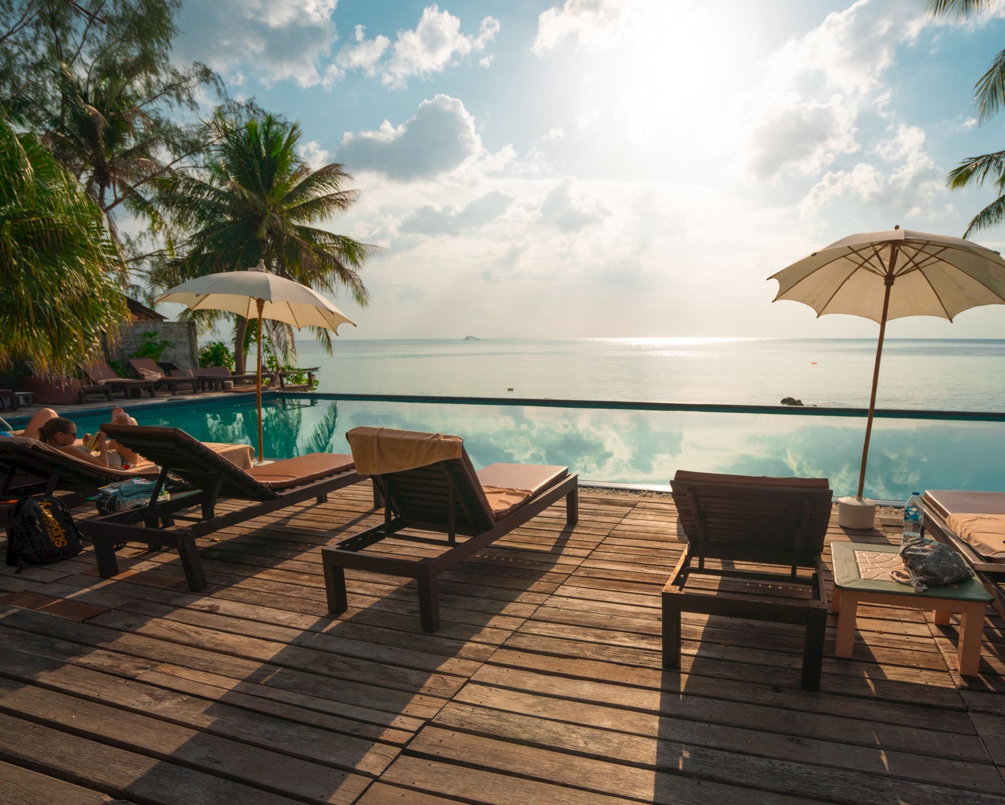 The image shows a tranquil beach scene with sun loungers, umbrellas, palm trees, and a calm sea under a cloudy sky, bathed in sunlight.
