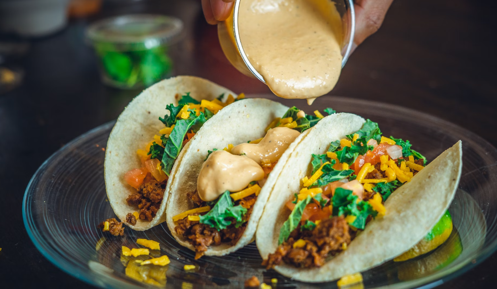 A hand is pouring sauce over three tacos filled with mixed greens, tomatoes, cheese, and ground meat, all served on a glass plate.