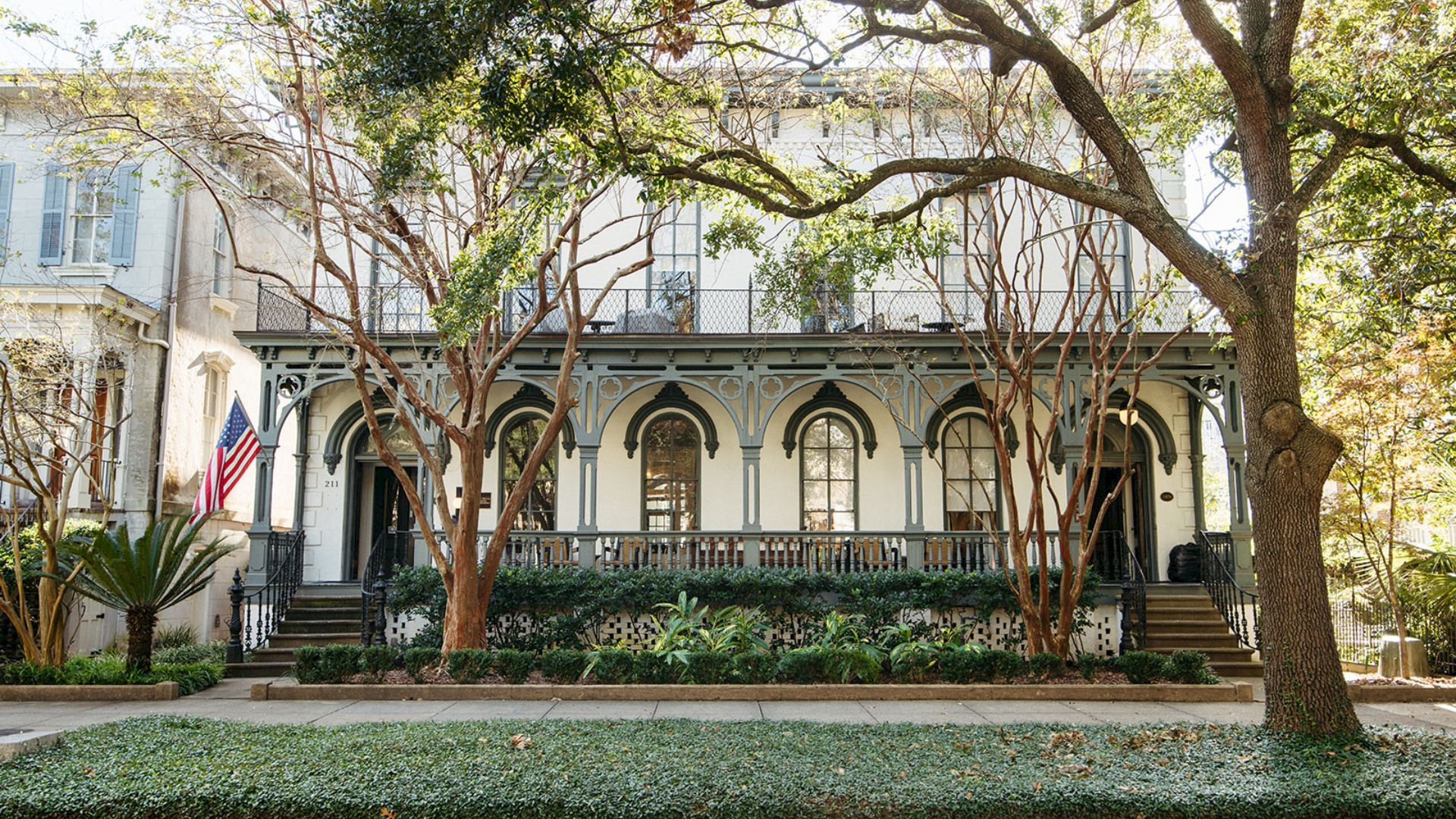 A white historic building with arched colonnade and a U.S. flag stands behind lush greenery and tall trees in front of it.
