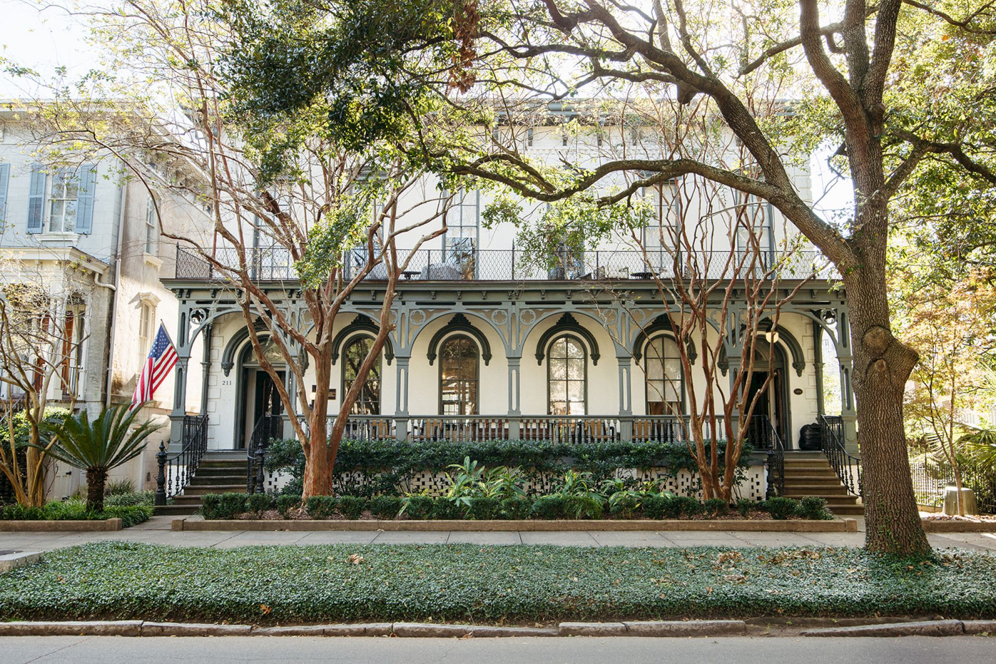 A historic house with ornate details, surrounded by trees, plants, and an American flag, creates a picturesque street view.