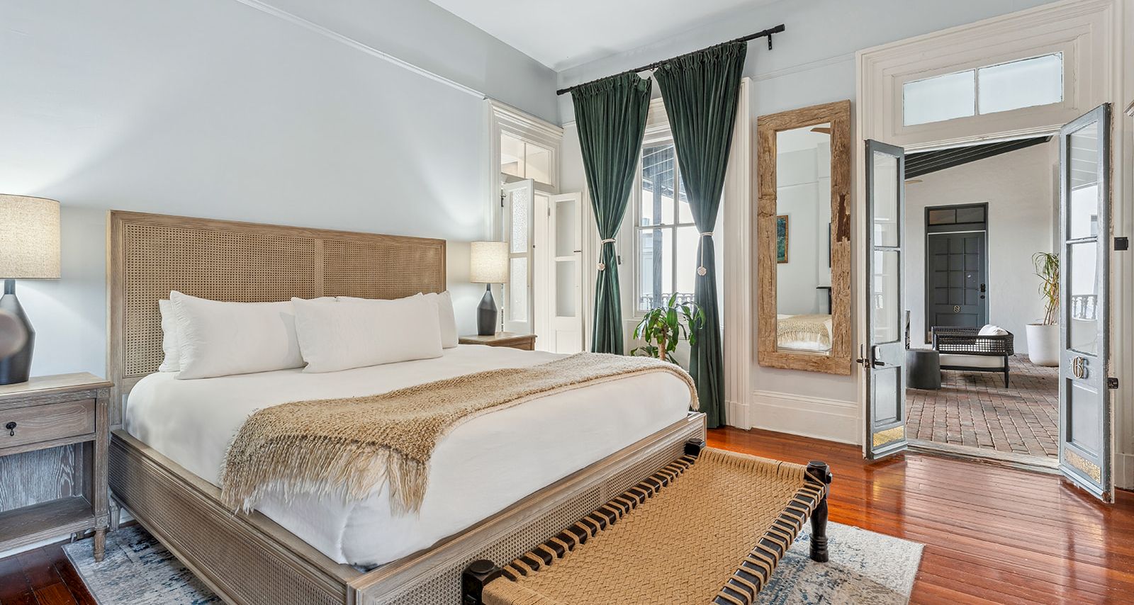 A bright hotel room with a king bed, nightstands, lamps, a bench at the foot, hardwood floors, and a doorway to another seating area.