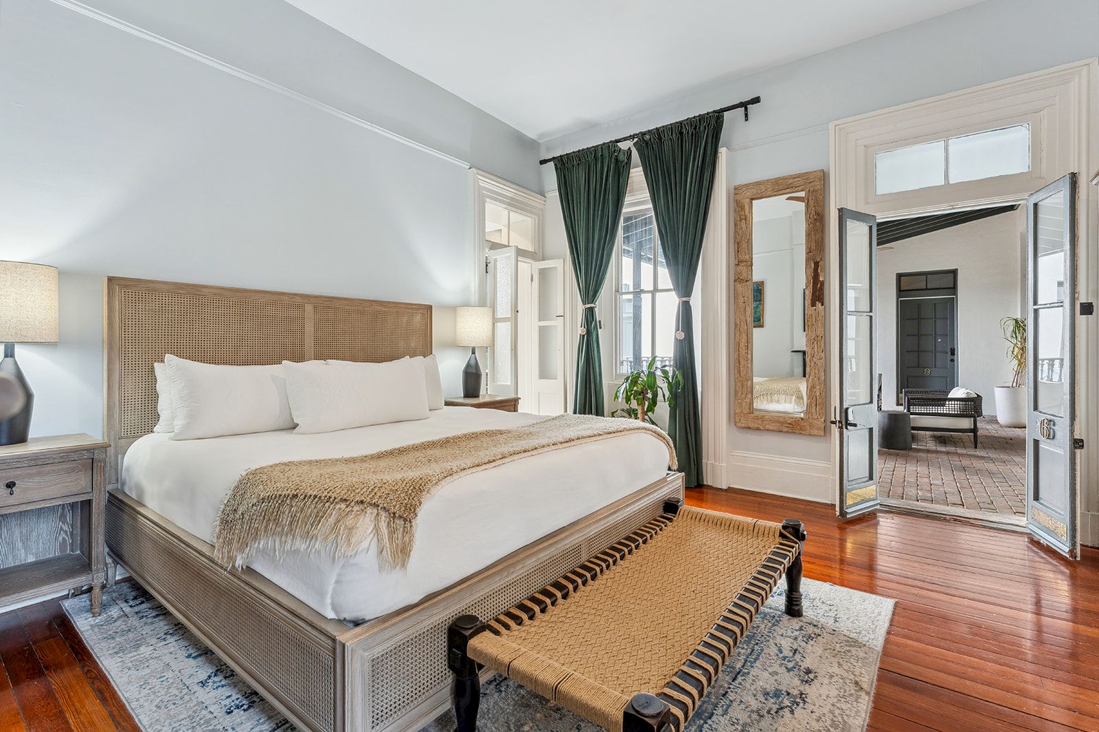 A bright hotel room with a king bed, nightstands, lamps, a bench at the foot, hardwood floors, and a doorway to another seating area.