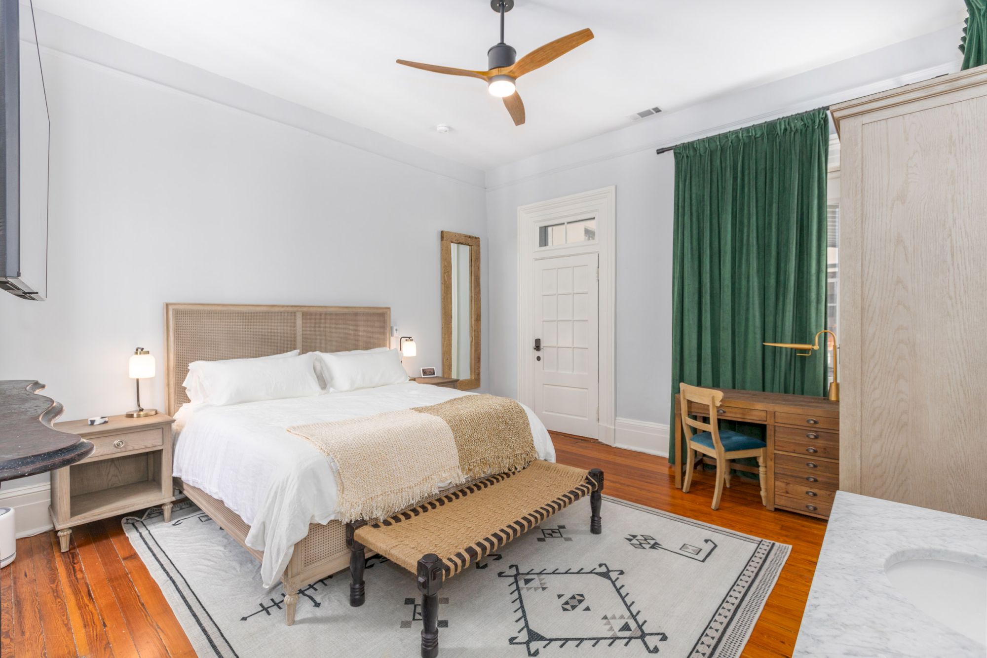 A modern bedroom with a bed, bench, desk, chair, rug, mirror, ceiling fan, and green curtains. The room has white walls and wooden floors.