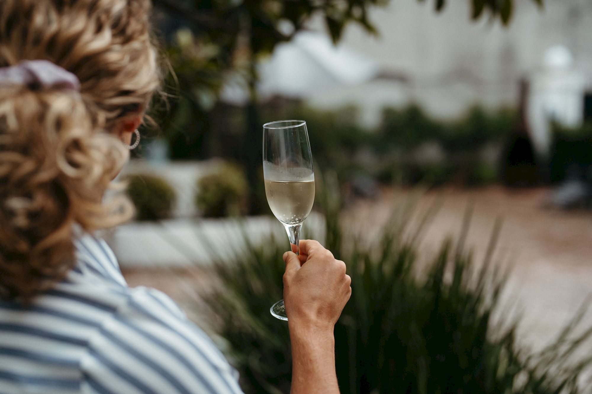 A person holding a glass of white wine, standing outdoors with greenery and blurred background. The focus is on the wine glass.
