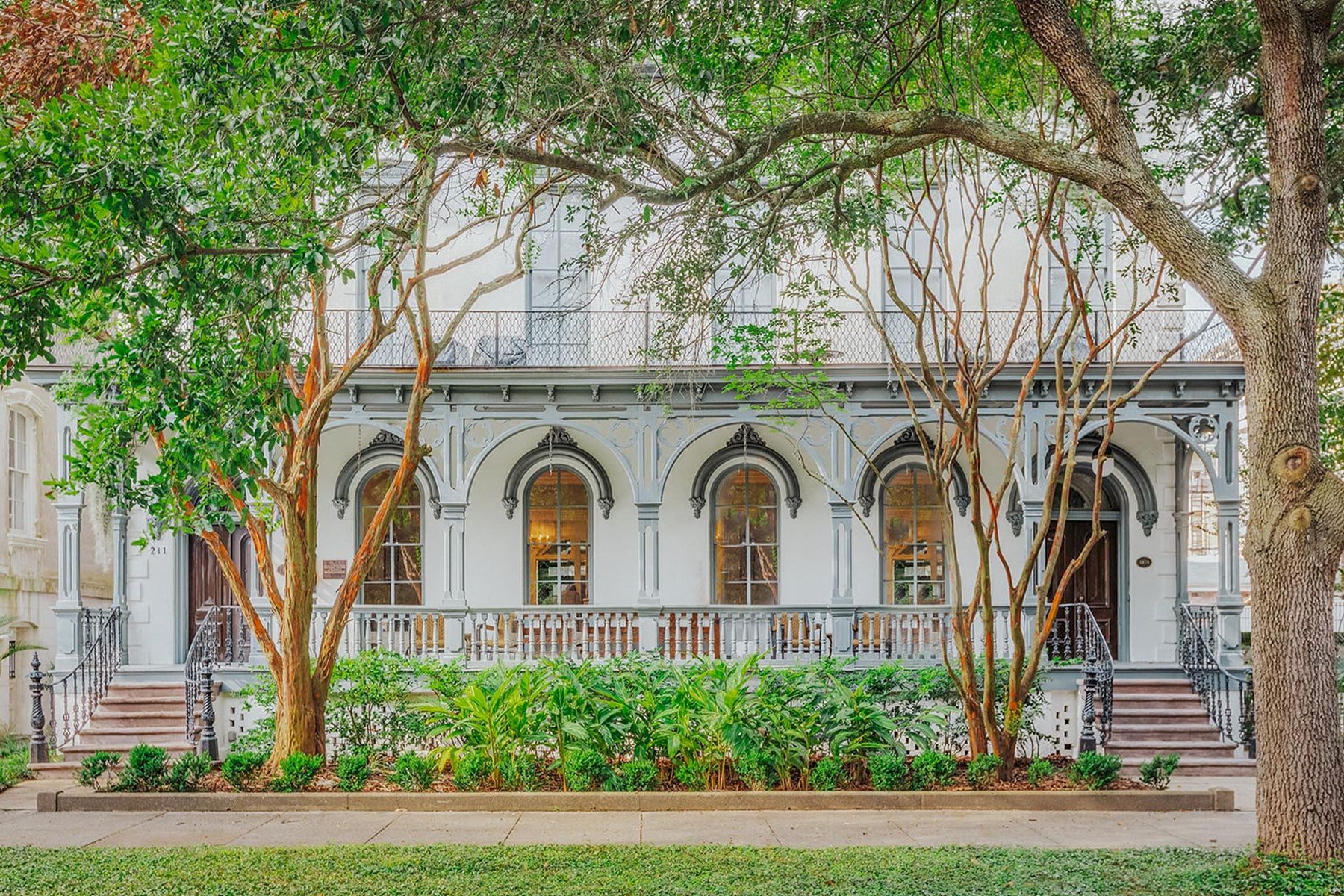 A white building with a balcony and stairs, surrounded by trees and bushes.