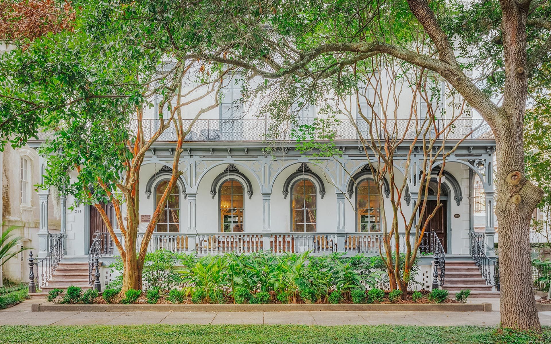 The image shows a charming two-story house with arched windows, a front porch, and lush greenery in the yard. Trees frame the view beautifully.