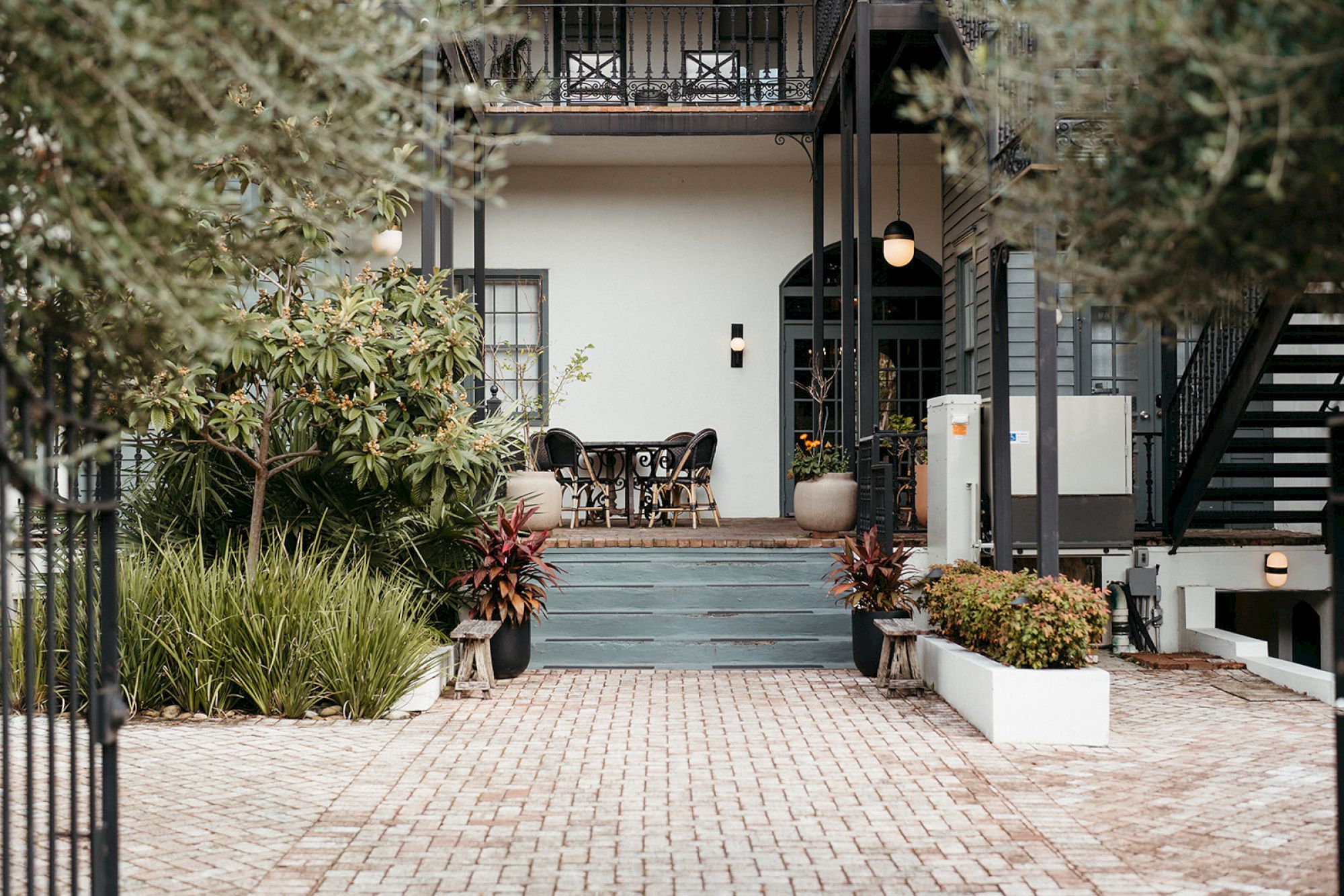 A charming building entrance with plants, a table, and chairs on a patio, surrounded by greenery and brick paving.