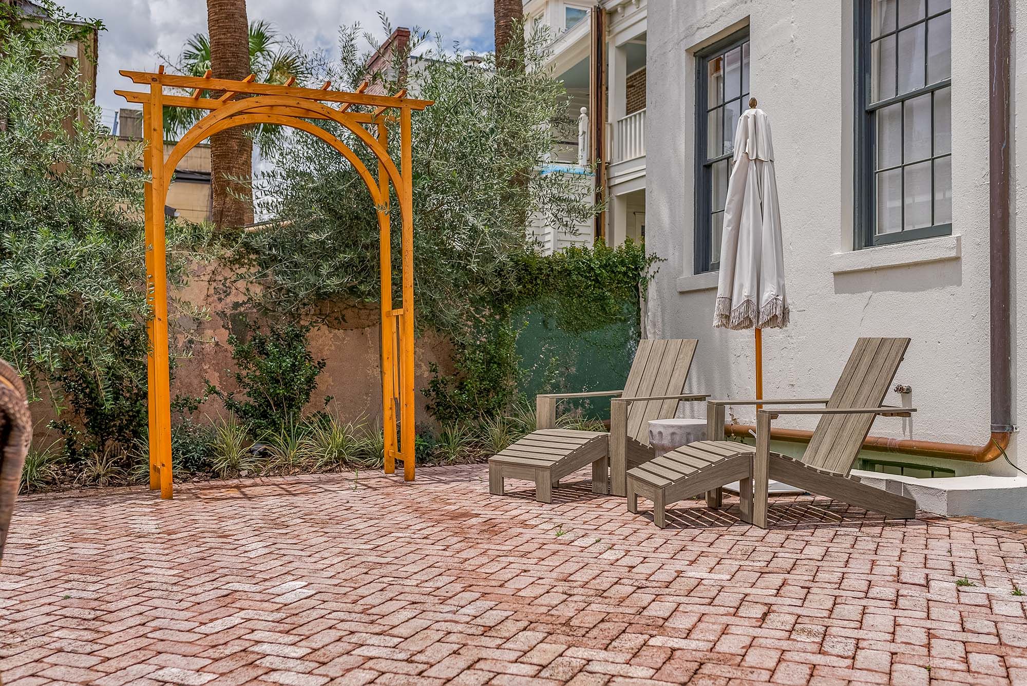 An outdoor patio with two wooden lounge chairs, an umbrella, and a yellow archway surrounded by greenery near a building.