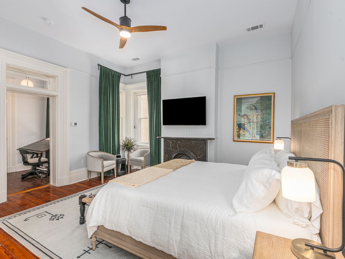 The image shows a modern bedroom with a bed, two chairs, a TV, and green curtains next to a large window. A ceiling fan is also visible.