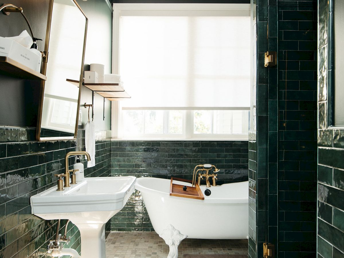 A bathroom with green tiled walls, a pedestal sink, a freestanding bathtub, and a window with a blind.