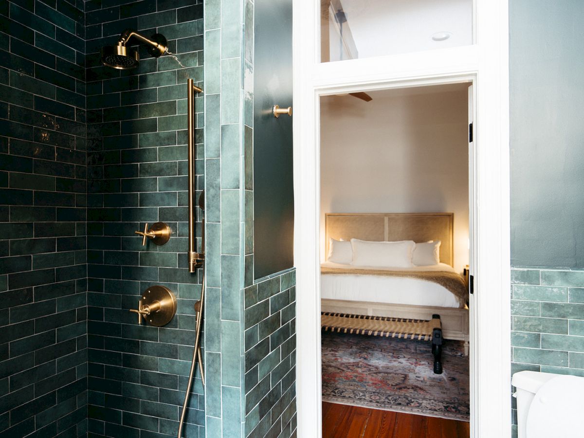 A stylish bathroom with dark green tiles and brass fixtures is adjacent to a cozy bedroom with a bed and rug.