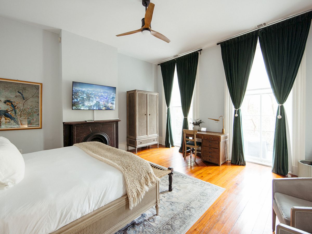 A modern bedroom with a bed, TV, desk, and tall curtains. The room features wooden floors, ceiling fan, and tasteful decor.