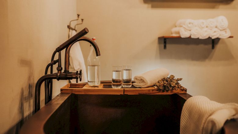 A cozy bathroom scene with a wooden bathtub tray holding water glasses, a towel, and a flower, with rolled towels on a shelf in the background.