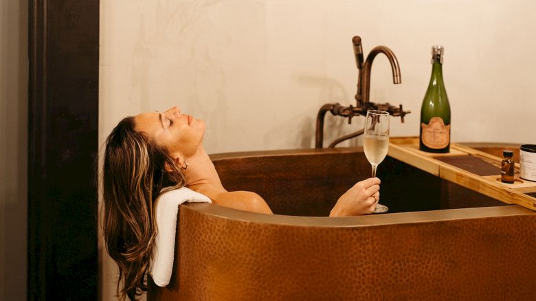 A person relaxes in a copper tub with a glass of wine in hand, while a champagne bottle and other items sit nearby on a wooden ledge.