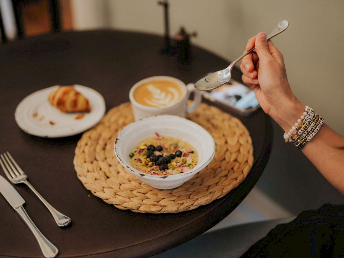 A person is having breakfast: oatmeal with berries, a croissant, and a latte on a table, with utensils on the side.