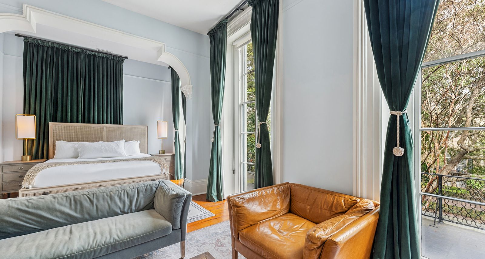 A stylish hotel room with a king bed, green drapes, a tan leather chair, a gray sofa, and large windows letting in natural light.