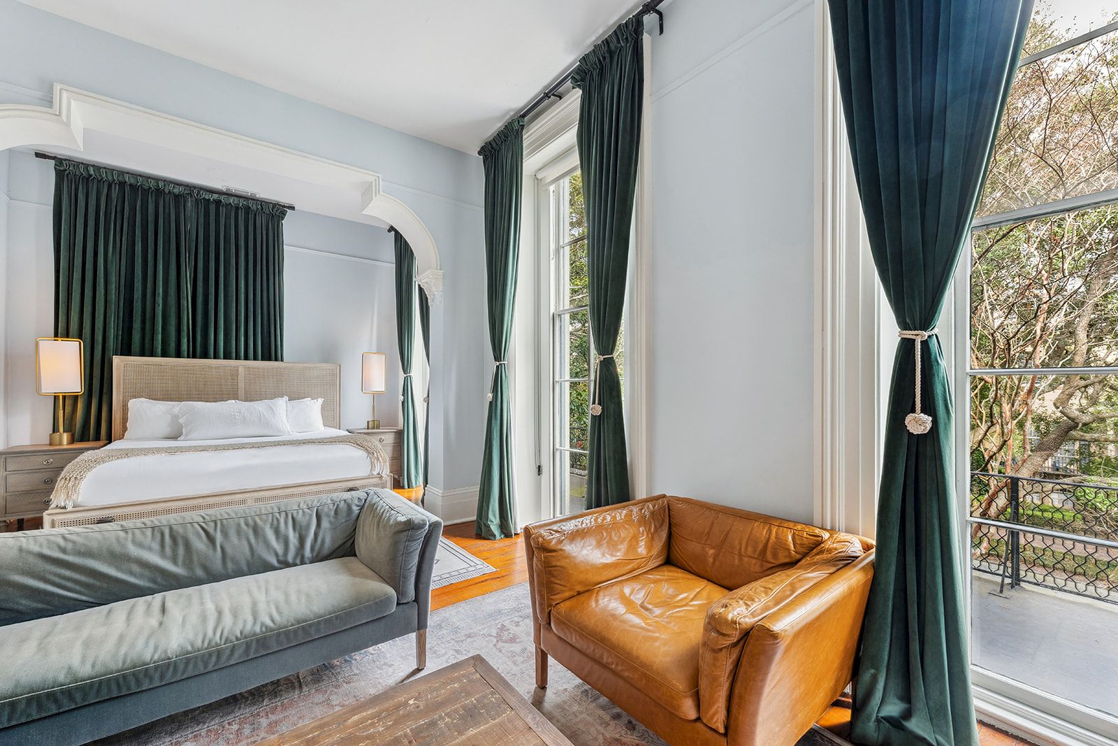 A stylish hotel room with a king bed, green drapes, a tan leather chair, a gray sofa, and large windows letting in natural light.