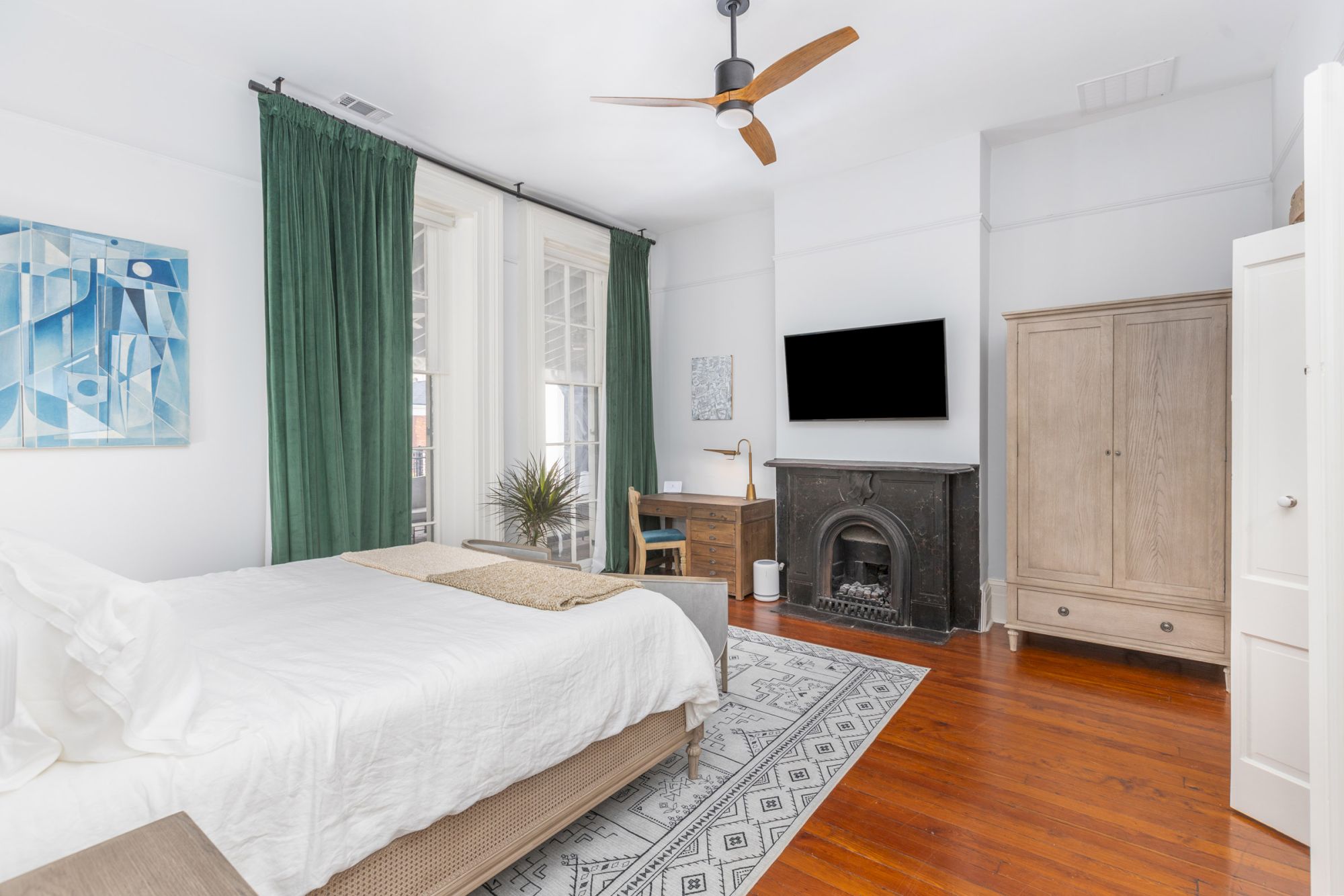 A tidy bedroom with a bed, green curtains, white linens, wooden floor, a TV above a fireplace, a desk, and a wardrobe.