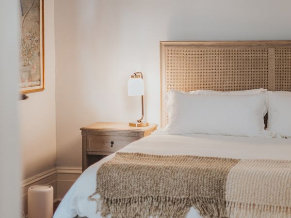 A cozy bedroom with a neatly made bed, a bedside table with a lamp, a wall-mounted artwork, and a woven bench at the foot of the bed.