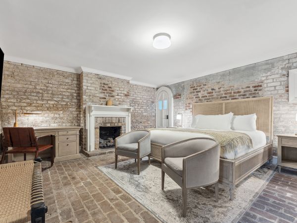 Luxurious brick-walled hotel room with a comfy king bed, two chairs, a fireplace, desk, and soft lighting for a cozy, rustic-modern vibe.