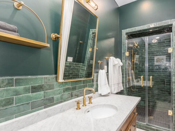A modern bathroom with green tiles, a marble countertop, gold fixtures, a large mirror, and a walk-in glass shower with towels hanging.
