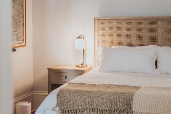 A cozy bedroom with a neatly made bed, a bedside table with a lamp, a wall-mounted artwork, and a woven bench at the foot of the bed.